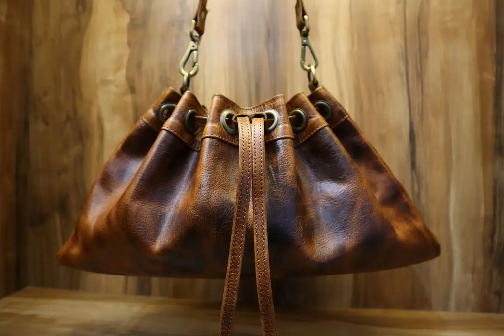 Brown leather bucket handbag with a drawstring closure, brass hardware, and shoulder strap. Vintage trunk-style purse.