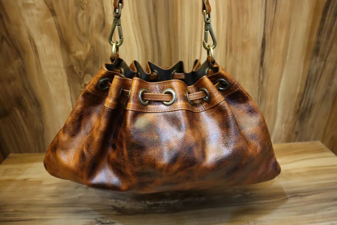 Brown leather bucket bag with brass grommets, drawstring closure, and shoulder strap. Vintage trunk-style aesthetic.