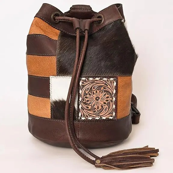 Brown leather bucket bag with western floral tooling, patchwork hair-on-hide panels, and a tassel drawstring closure.