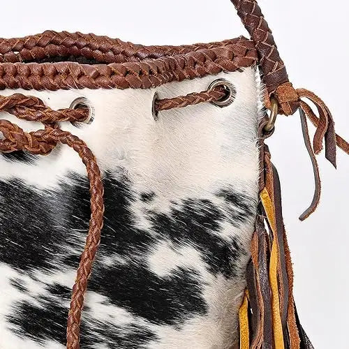 Cow print faux fur bucket bag with brown braided leather trim, tassel fringe, and drawstring closure for women.