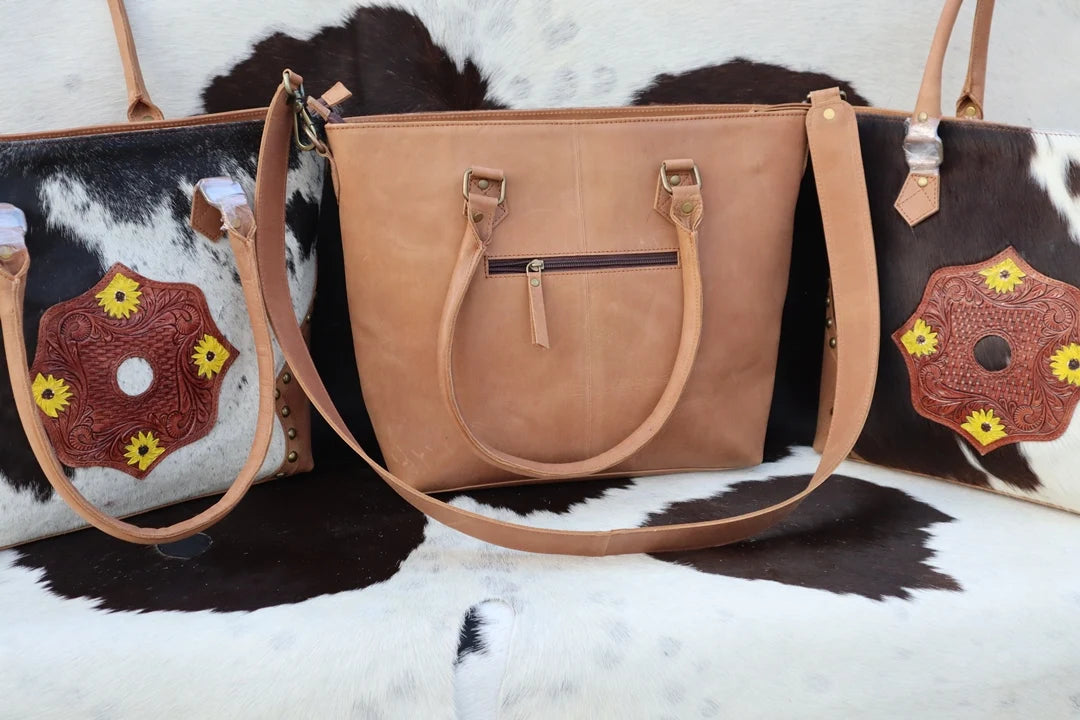 RABADA Western leather tote with hair-on hide, sunflower tooling, zipper closure, and dual carry straps on cowhide rug.