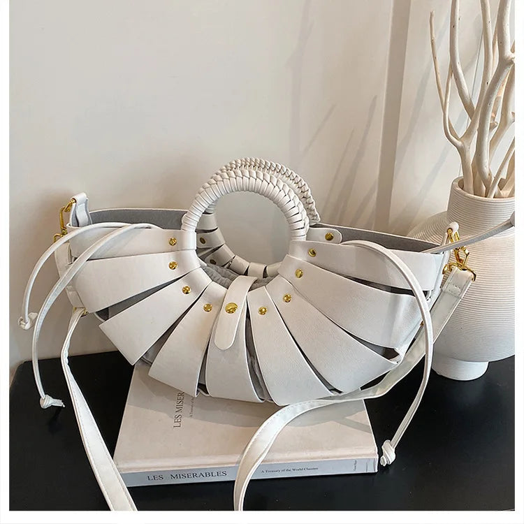 White half-moon trunk bag with hollow-out leather straps, round braided handle, and gold hardware shoulder strap.