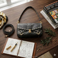 Black calf leather shoulder bag with gold studs, flap closure, and trunk-style detailing on a wooden desk.