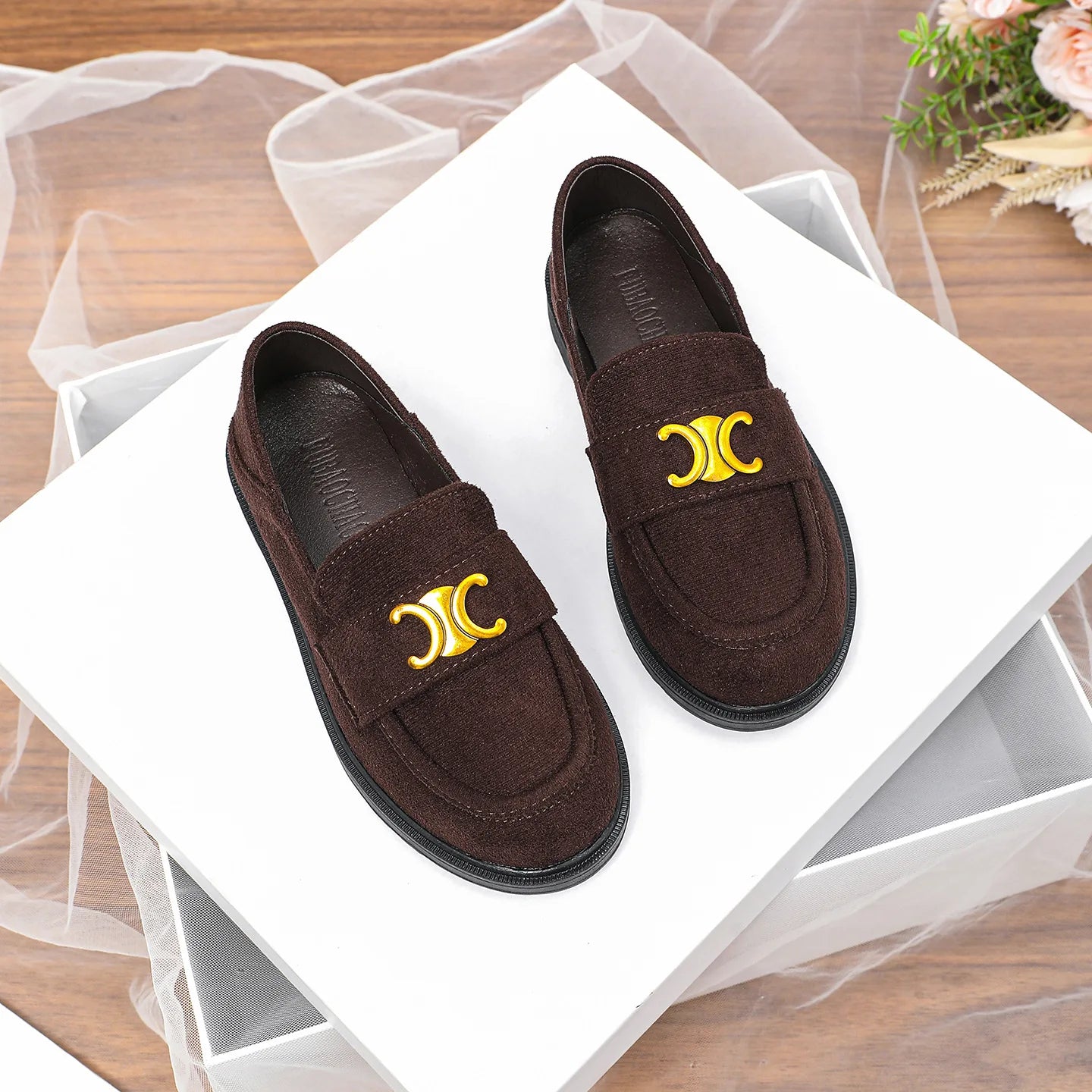 Luxury dark brown suede loafers for women with gold logo embellishment and flat black rubber sole on white display box.