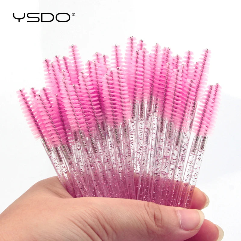 Hand holding a bundle of disposable pink eyelash spoolie brushes with glittery transparent plastic handles.