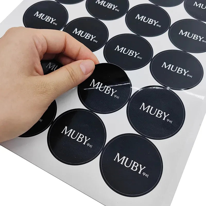 Hand holding round dark MUBY logo stickers on a backing sheet