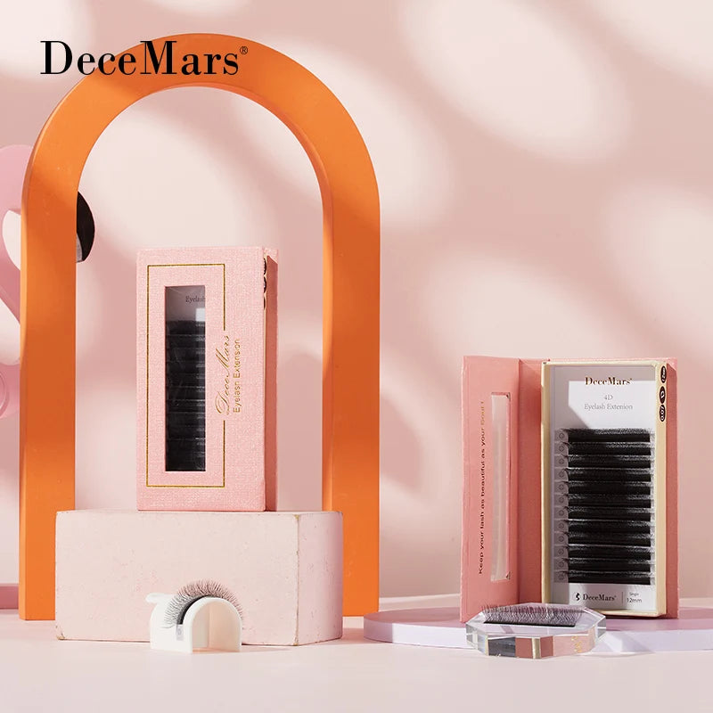Two pink boxes of DeceMars 4D W-shaped eyelash extensions (0.07mm) displayed with loose lash strips on a pink set.