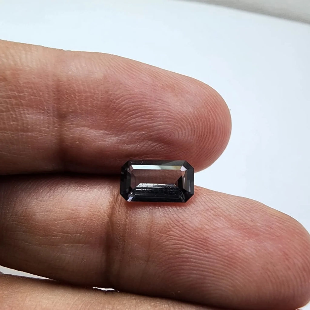 Small dark gray emerald-cut spinel gemstone held on fingertip