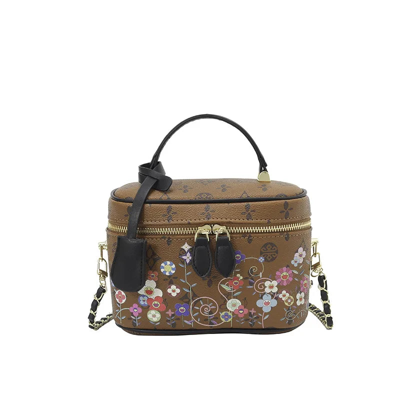 Brown leather vanity trunk bag with floral monogram, gold zipper, black handle, and detachable chain shoulder strap.