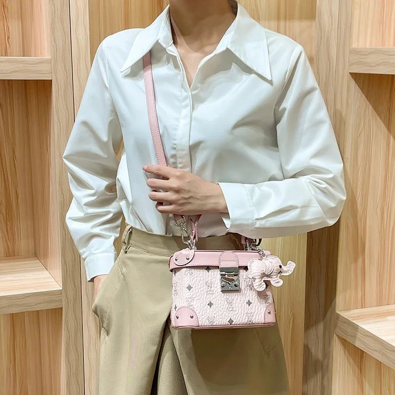 Minimalist pink cowhide leather trunk handbag with embossed logo pattern, metallic clasp, and elephant bag charm.