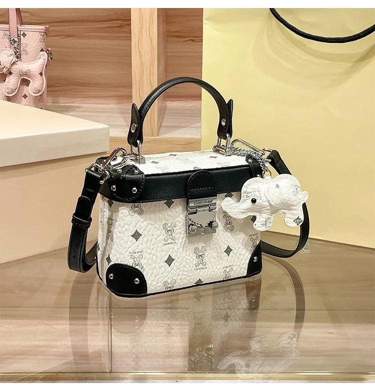 White and black cowhide trunk handbag with minimalist monogram print, metal lock clasp, and cute elephant charm.