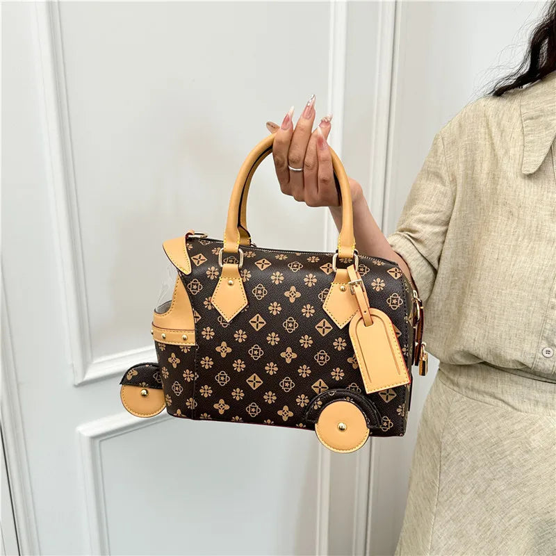 Brown patterned PU leather trunk handbag with beige trim, top handle, luggage tag, and unique tricycle wheel accents.