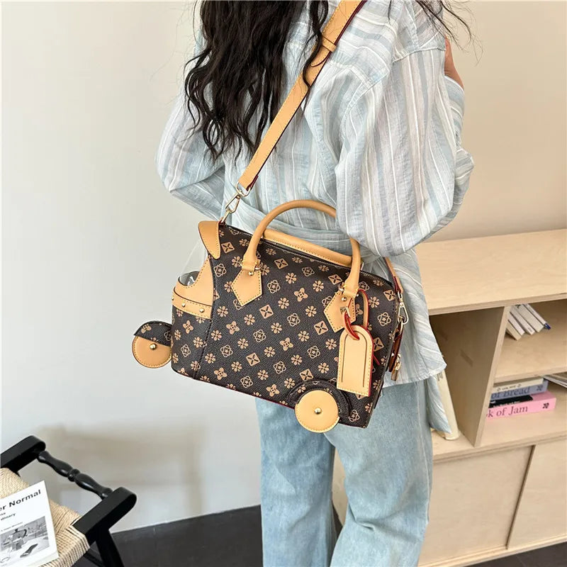 Brown patterned PU leather trunk handbag with tricycle wheels, tan trim, and detachable shoulder strap.