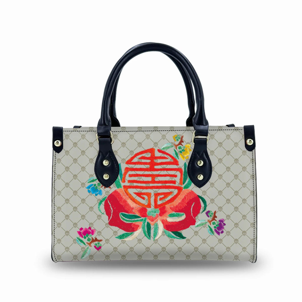 Beige faux leather trunk handbag with black handles, skull pattern, and colorful embroidered floral design.
