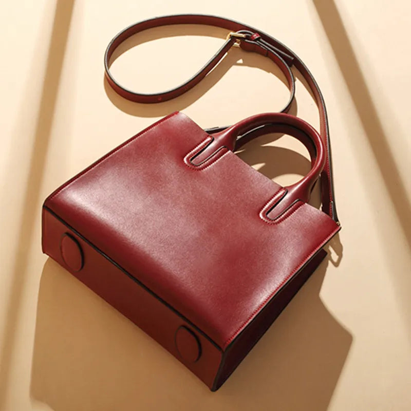 Burgundy leather trunk-style handbag with top handles, adjustable shoulder strap, and minimalist structured design.