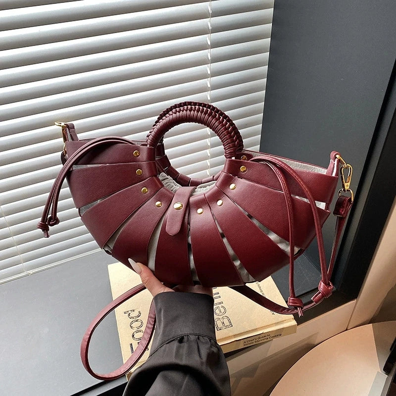 Burgundy faux leather half-moon crossbody bag with cutout design, circular braided handle, and gold stud details.