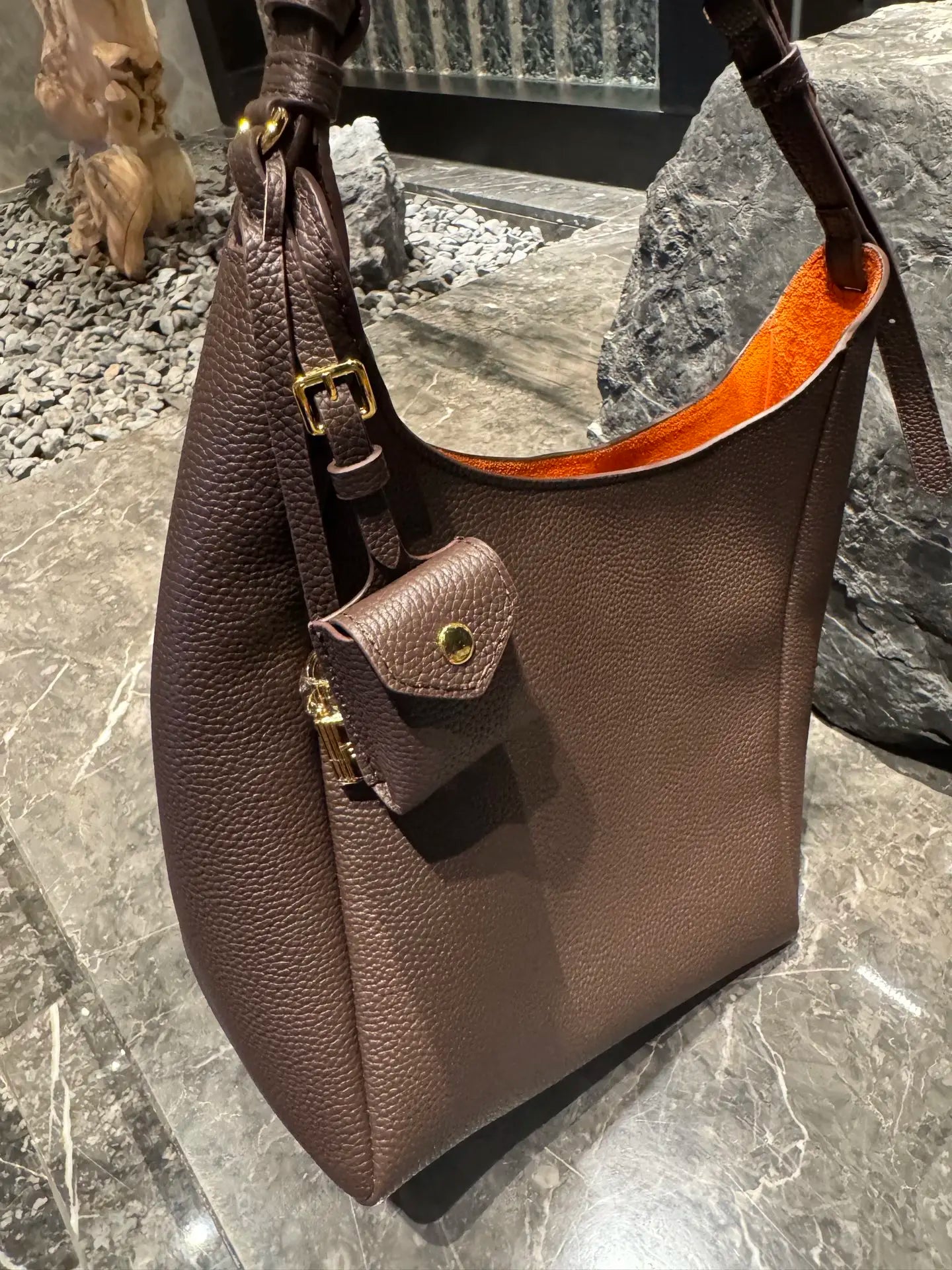 Brown pebble-grain leather hobo bag with gold hardware, adjustable strap, and contrast orange interior. Luxury trunk.
