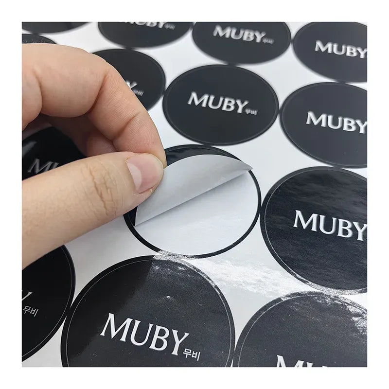 Close-up of a hand peeling a circular dark MUBY logo label sticker