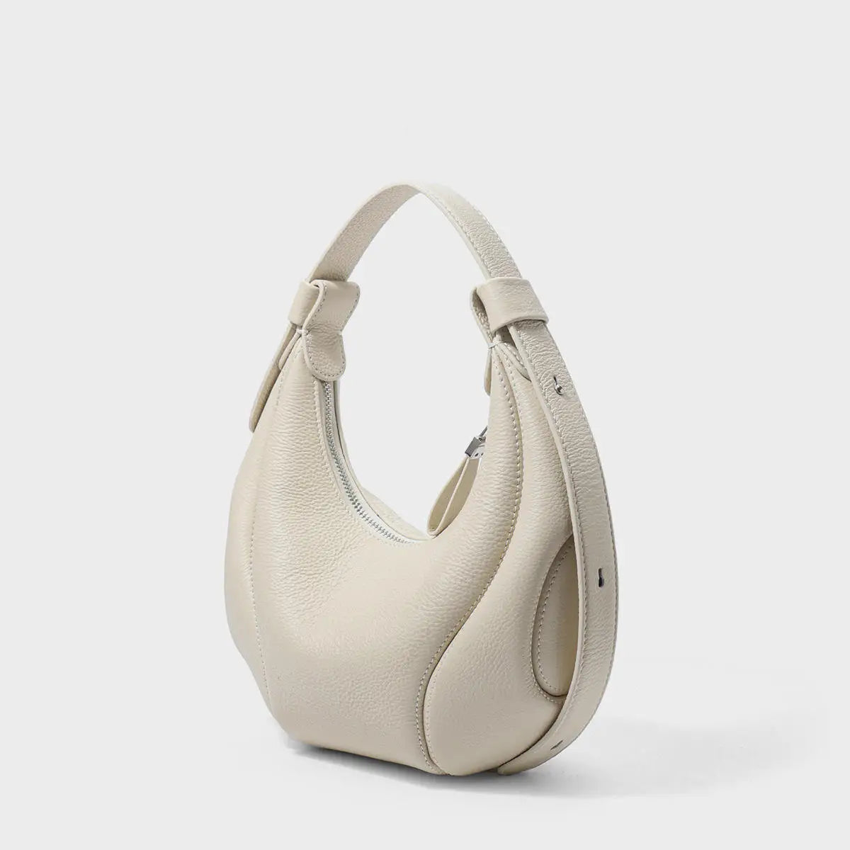Cream pebble leather half-moon shoulder bag with zip closure and adjustable strap, elegant designer trunk style.