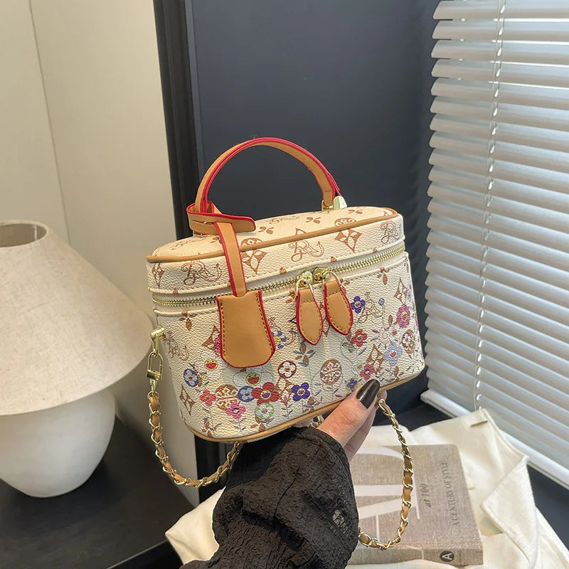 Cream genuine leather trunk bag with colorful floral print, red trim, gold chain strap, and zipper top handle.