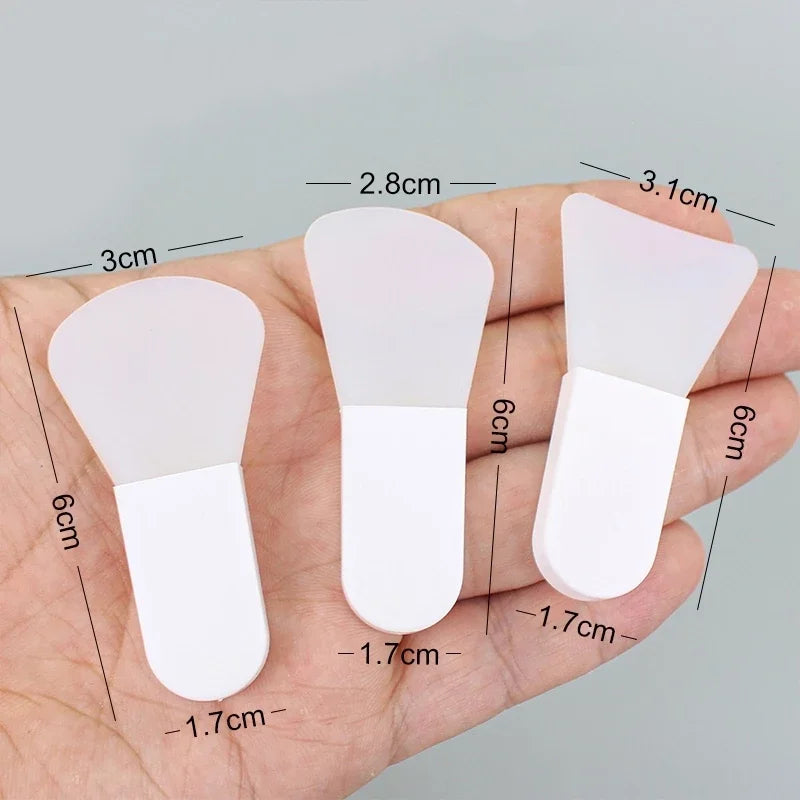 Three small, white silicone facial mask brushes with varying head shapes, held in a hand, showing size dimensions.