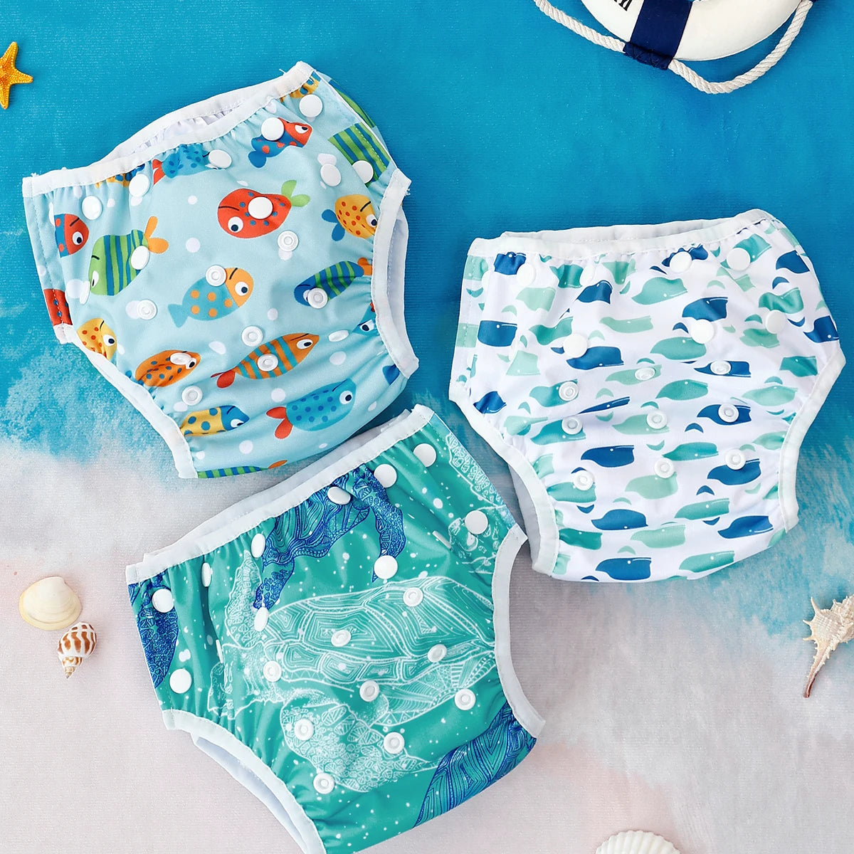 Three reusable baby swim diapers with ocean fish, whale, and sea turtle prints on a beach background.