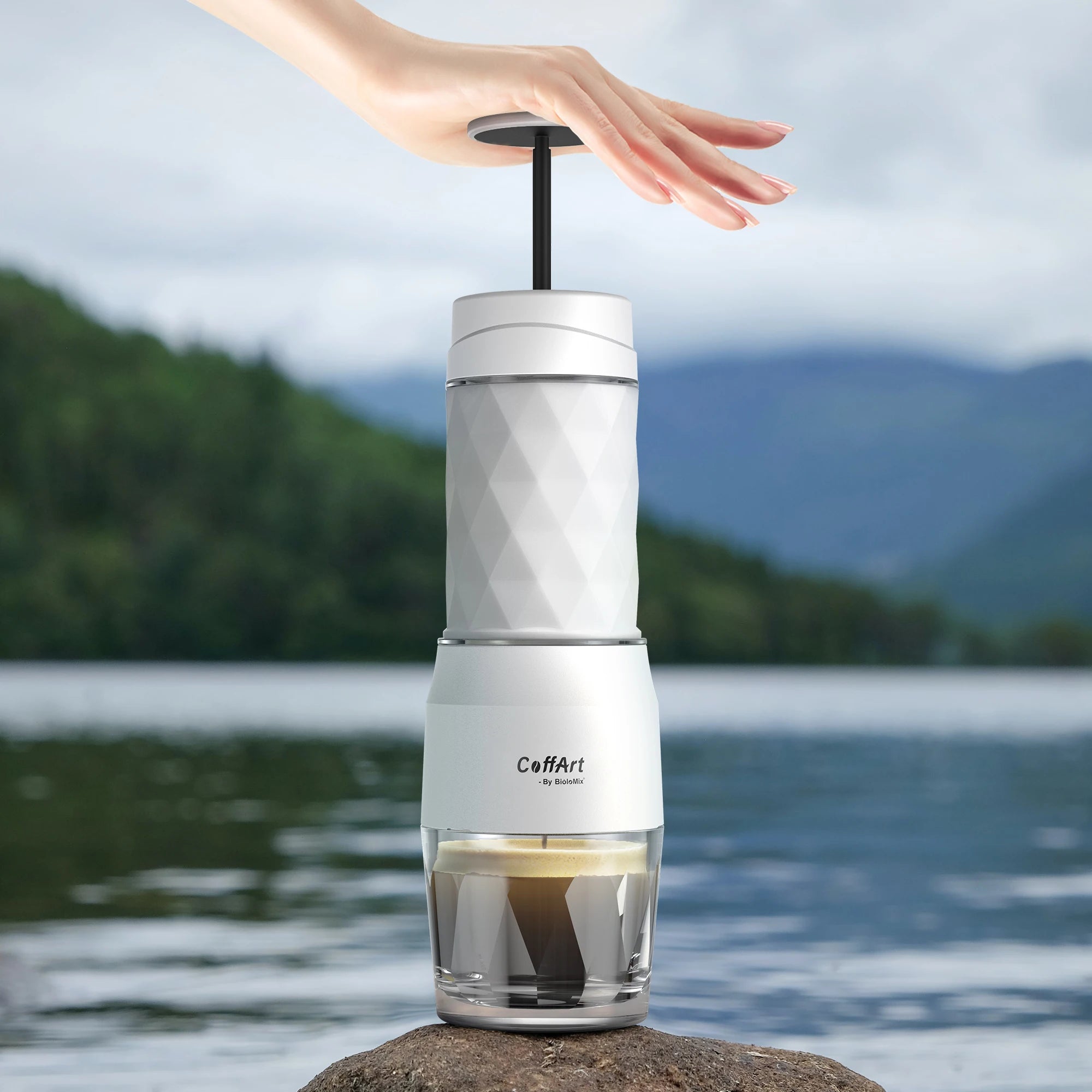 CoffArt by BioloMix portable white manual espresso maker with diamond-textured body brewing coffee on a lakeside rock.
