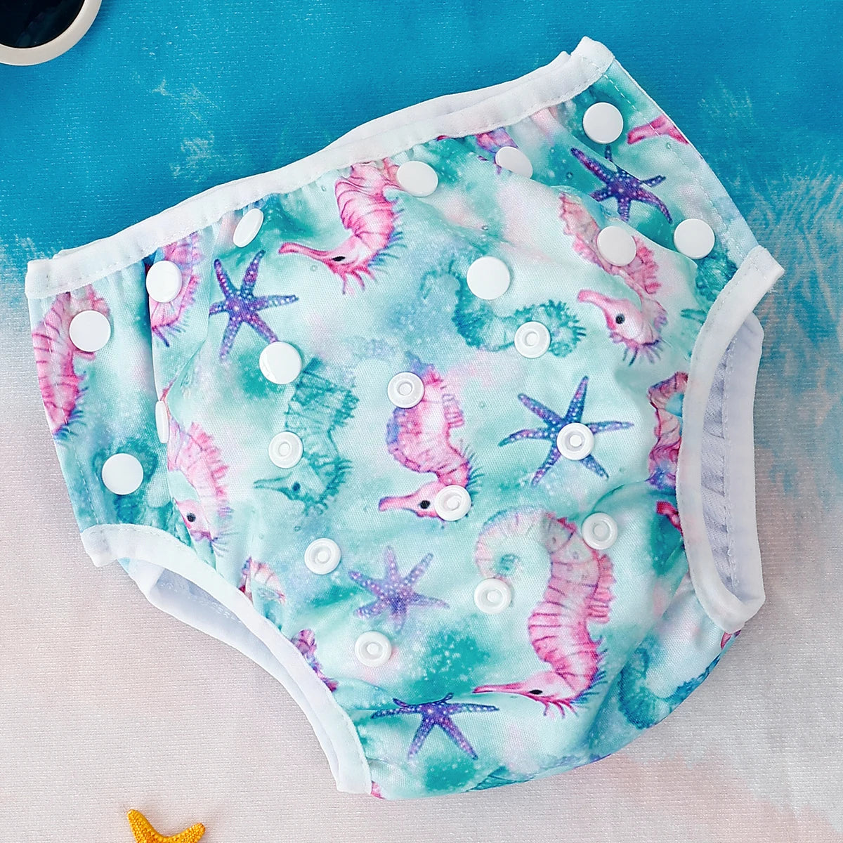 Reusable baby swim diaper with white snap closures, featuring a teal sea horse and purple starfish print.