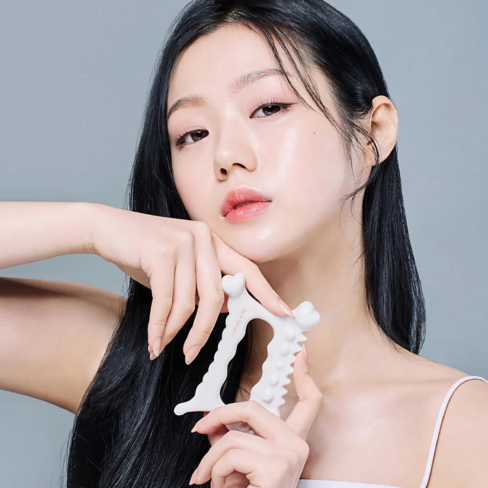 A woman holds a white, U-shaped ceramic gua sha facial massager with textured, spiked edges.
