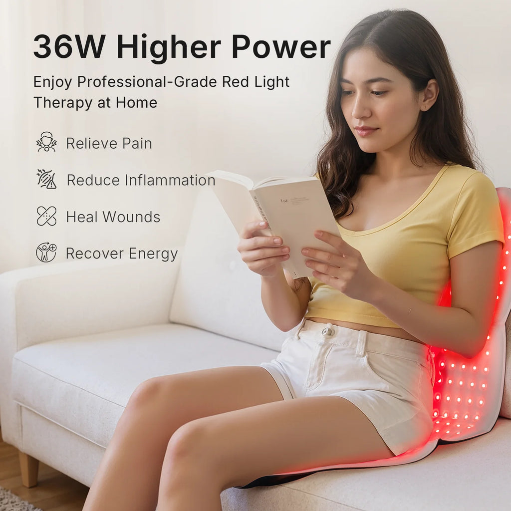 A woman sits on a sofa reading while using a flexible red light therapy mat against her back.