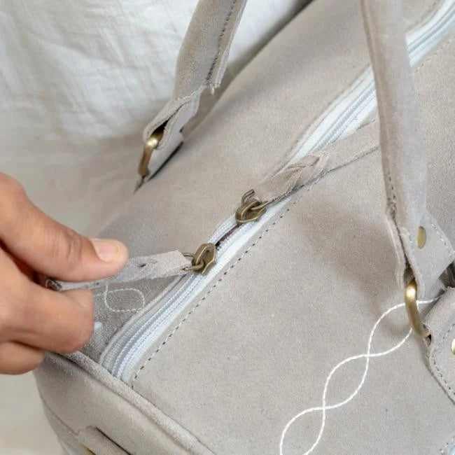 Handmade grey suede travel duffel bag with white boot stitch detail, brass hardware, and braided leather zip pulls.
