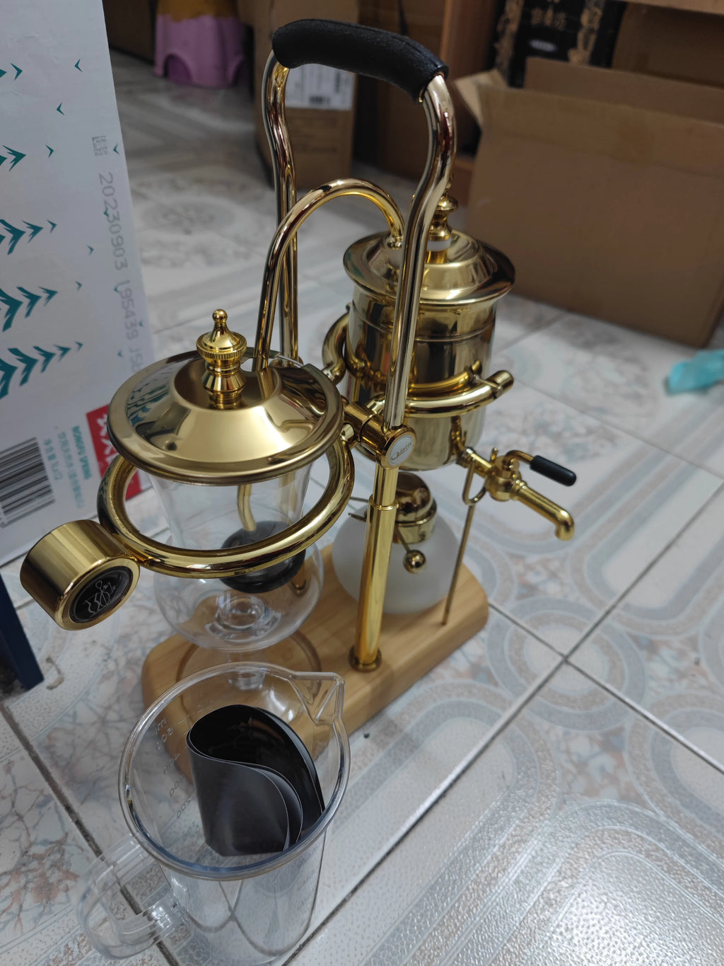 Gold balancing siphon coffee maker with glass chambers, metal finish, and wooden base stand for manual brewing.