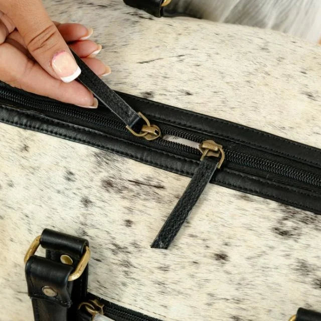 White and black spotted cow hair-on-hide weekender duffel bag with black leather trim and brass zipper hardware.
