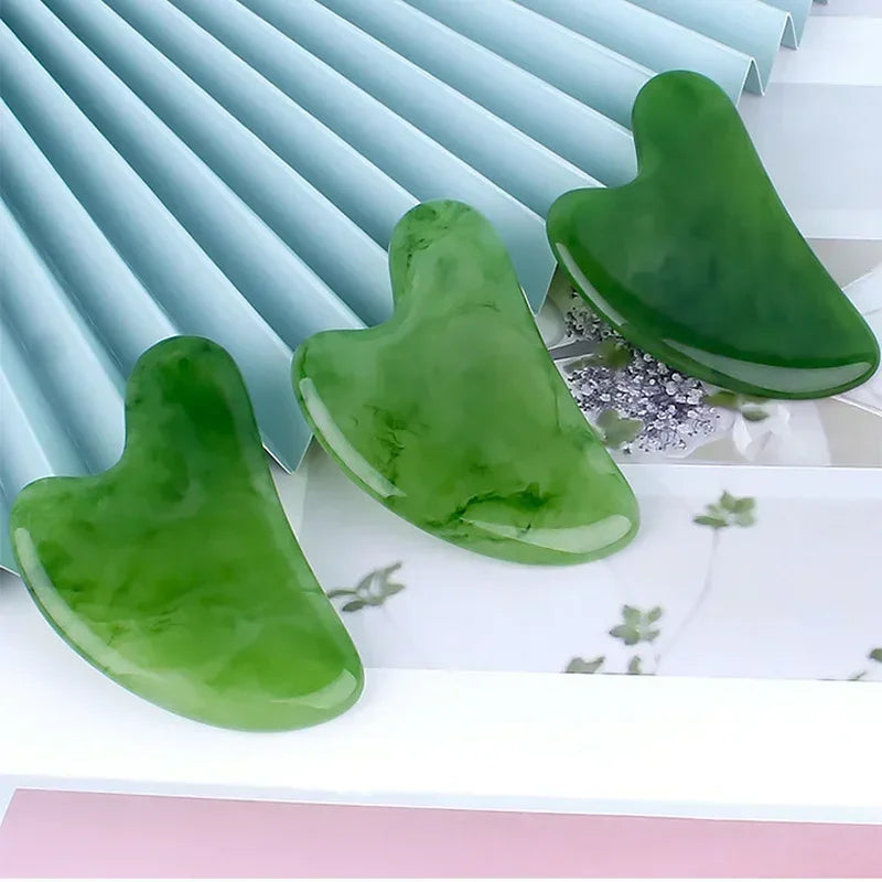 Three heart-shaped, polished green resin Gua Sha facial massage tools arranged on a decorative surface.