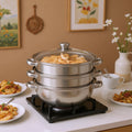 3-tier stainless steel steam cooker pot with glass lid and side handles, on black burner.