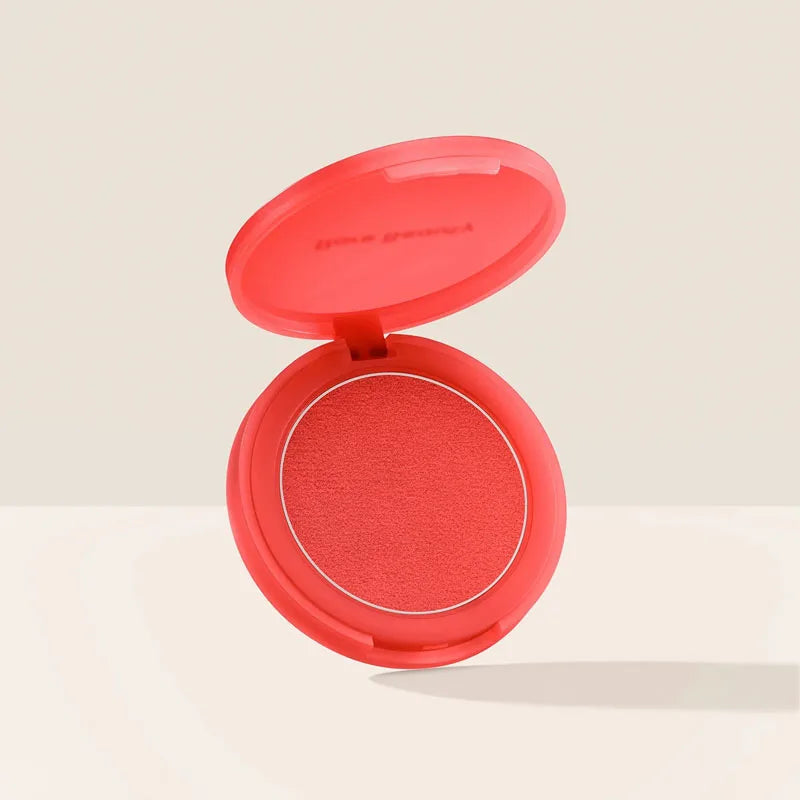 Rare Beauty powder blush in a vibrant coral shade with a smooth, matte mud-like texture in an open compact case.