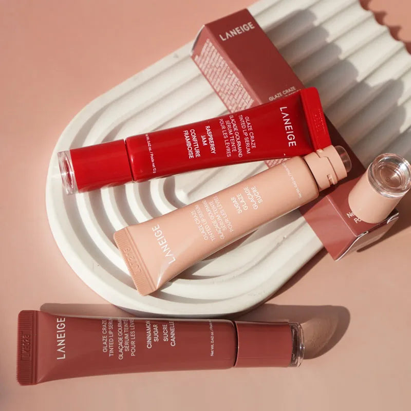 Laneige Glazey Tinted Lip Serum tubes in Raspberry, Sugar, and Cinnamon shades on a white tray. Hydrating gloss makeup.