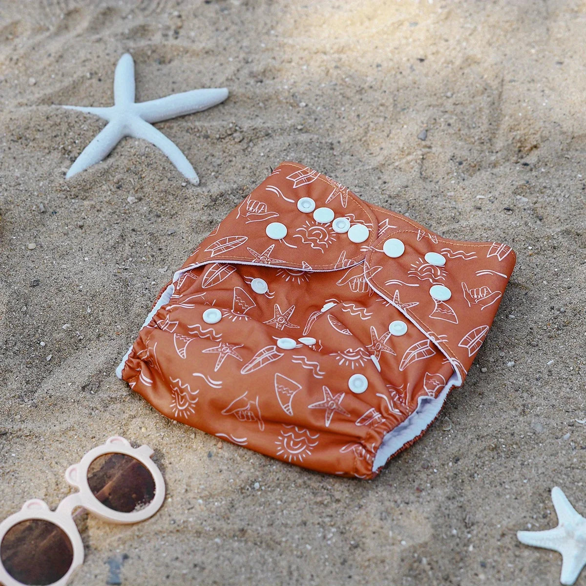 Reusable cloth diaper in rust orange with white summer surf print and snaps, displayed on beach sand with starfish.