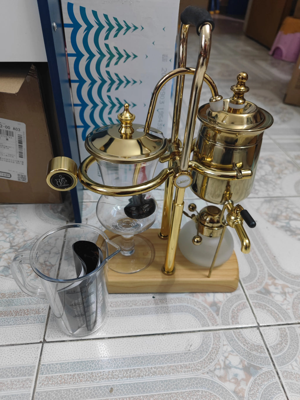 Gold-tone Balmain balanced siphon coffee maker with glass chambers, wooden base, and clear plastic measuring cup.