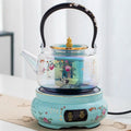 Decorative glass tea maker with crane and peach art on an electric turquoise ceramic heating base.