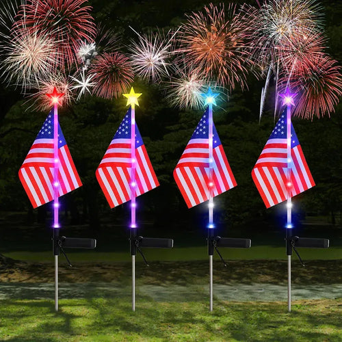 Four LED American flag solar garden stakes with glowing star toppers stand upright against a dark, firework-lit background.