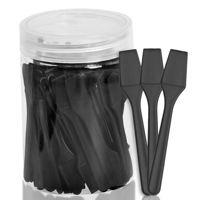 Clear jar filled with 100 small, black plastic cosmetic spatulas for applying face cream or makeup.