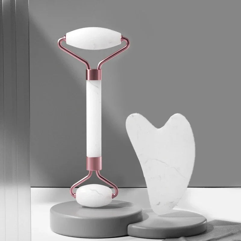 White marble face roller with rose gold accents and a matching heart-shaped gua sha tool on a grey circular base.