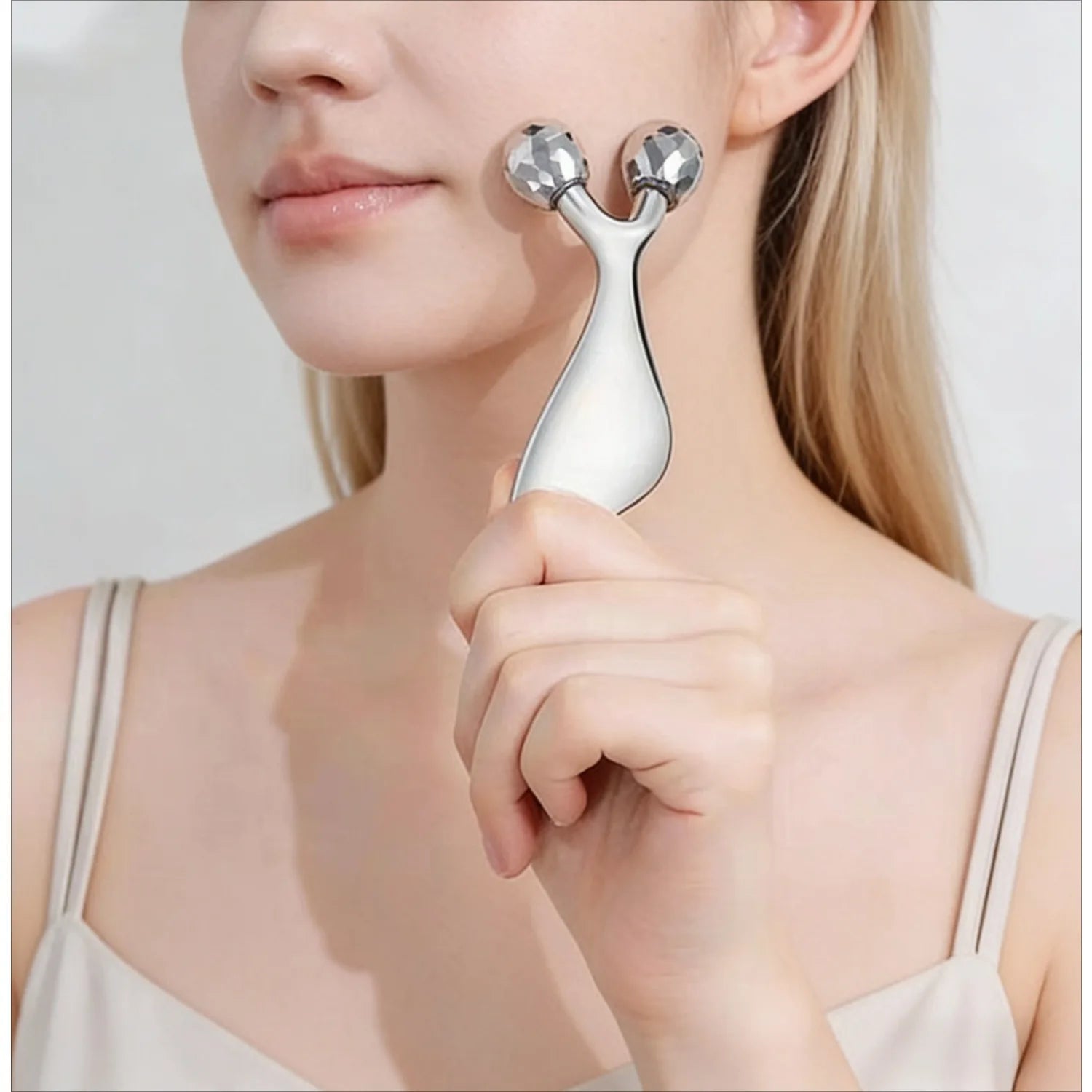 A woman holds a silver, Y-shaped facial massager with two textured, spherical roller heads against her cheek.