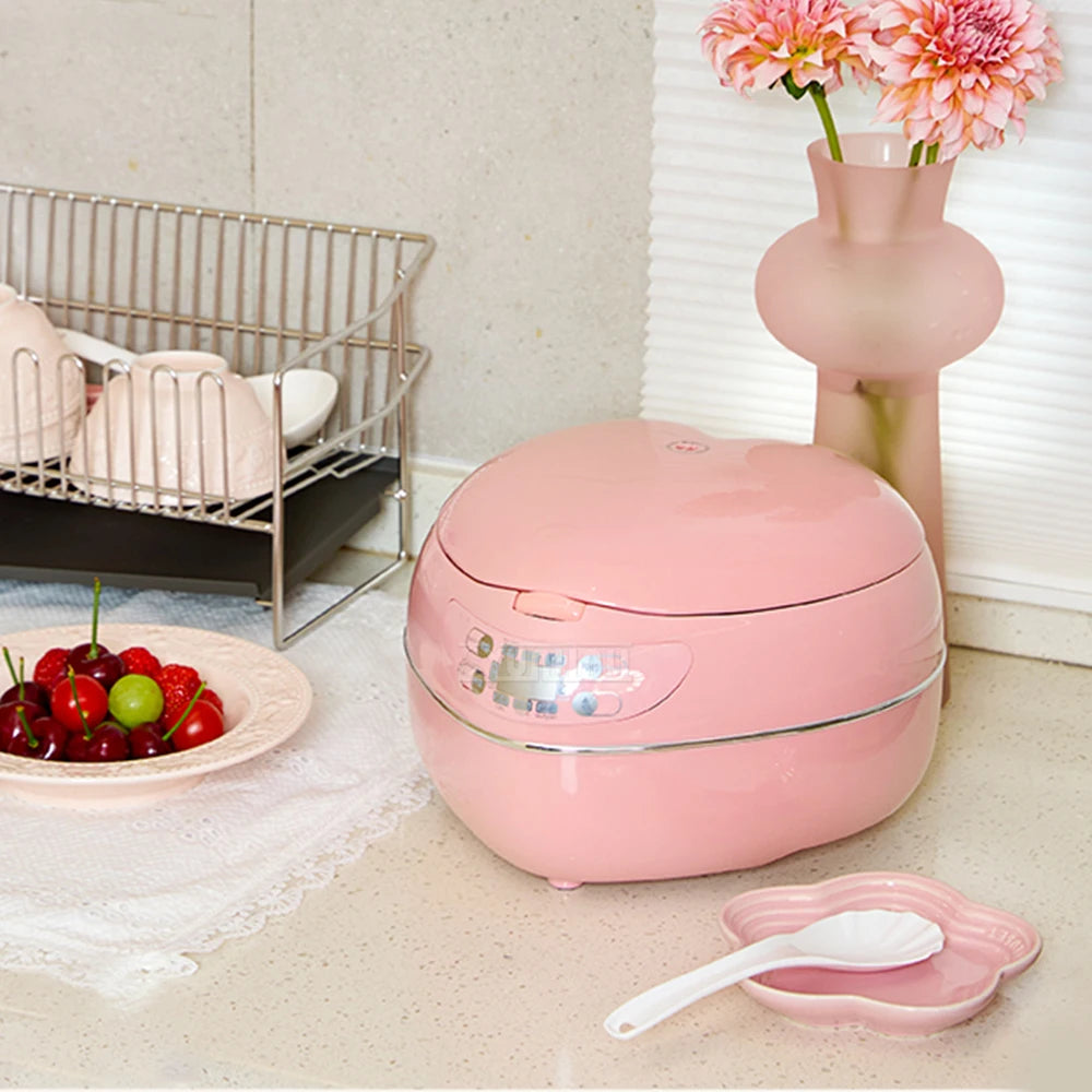 Pink heart-shaped mini rice cooker with lid closed beside accessories and dishes.
