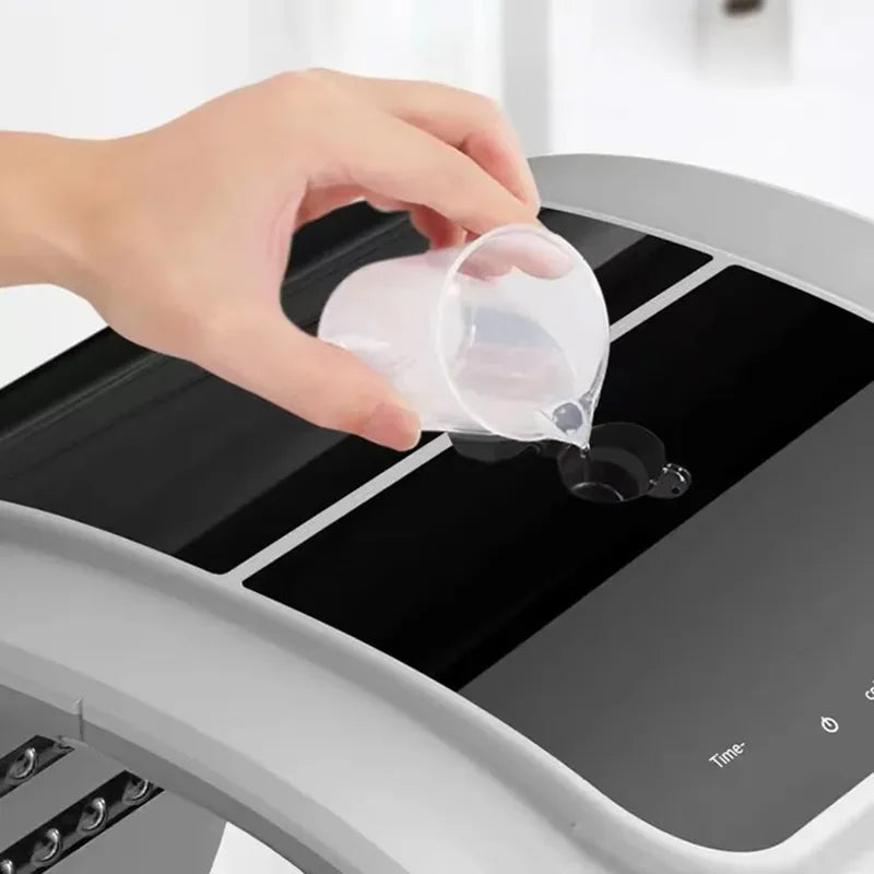 A hand pouring water from a small plastic cup into the reservoir of a grey and black LED facial beauty machine.
