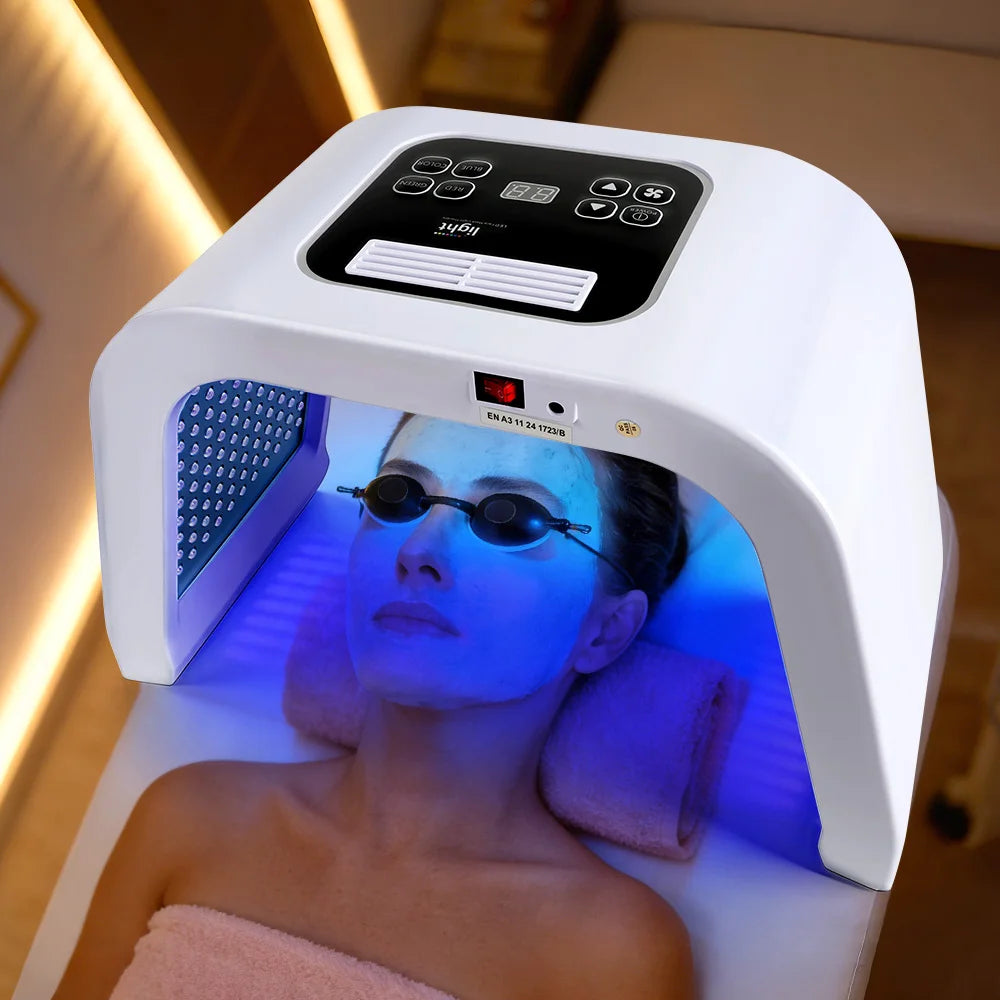 Person lying under a white, arched 7-color LED facial therapy device while wearing protective black goggles.