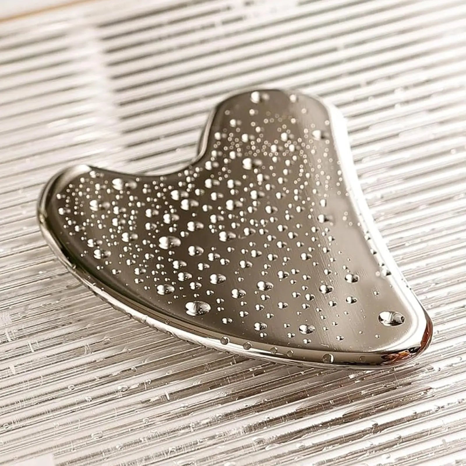 Heart-shaped stainless steel gua sha facial tool with water droplets, resting on a reflective, ribbed silver surface.