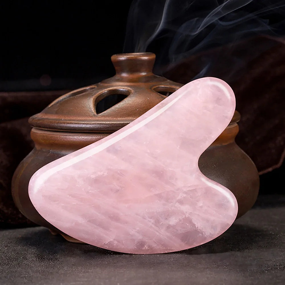 A heart-shaped, translucent pink resin gua sha massage tool placed in front of a smoking incense burner.