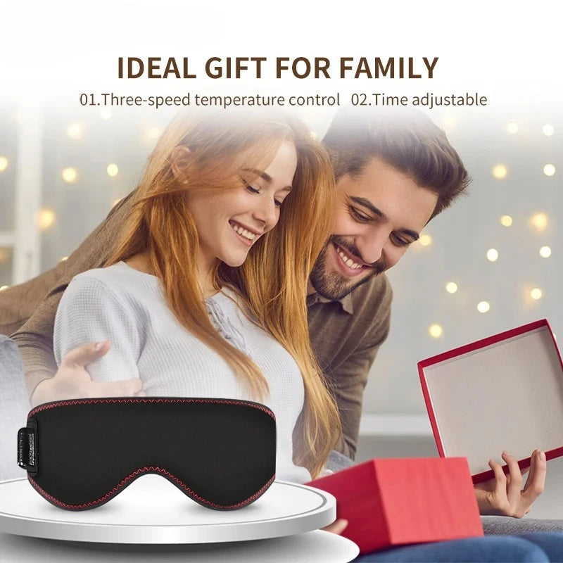 Black ergonomic heated eye mask with red stitching on a white display stand, with a blurred couple gifting in the back.